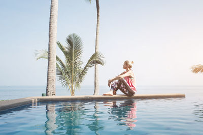 Side view of mid adult woman sitting at infinity pool against sea