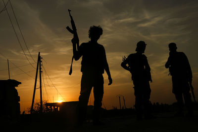 Silhouette men with guns standing against orange sky during sunset