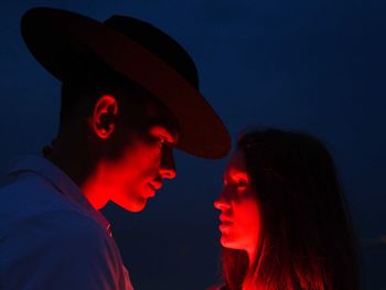 Side view of young couple standing against dark sky lit up by red neon light