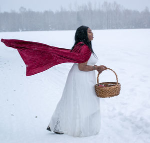 Side view of woman with red cape holding basket on landscape during winter