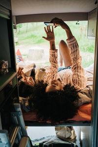 Young multiracial girlfriends lying together in camper van and taking selfie on smartphone while chilling and enjoying summer holidays in nature