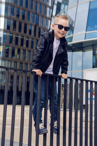 Baby boy in sunglasses and black leather jacket smiling on the background of the glass building