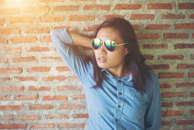 Young woman wearing sunglasses while standing with hands behind head against brick wall
