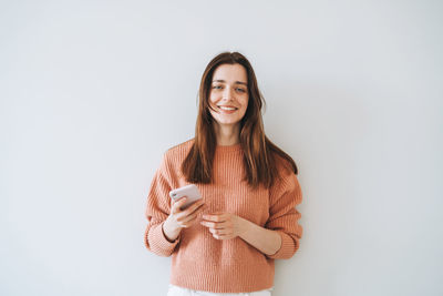 Portrait of smiling woman in casual knitted sweater using mobile phone in hand on grey background