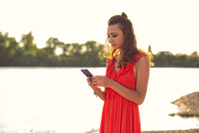 Young woman looking away while standing on smart phone against sky