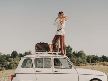 Full body side view of stylish female hippie in boho clothes standing on white old timer automobile with luggage during trip in nature