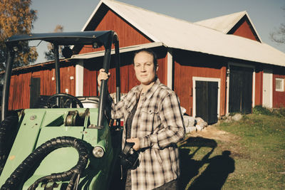 Portrait of confident female farmer standing by agricultural vehicle against barn