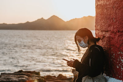 Side view of woman wearing mask using phone against sea