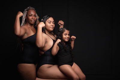 Group of sporty multiethnic multigenerational females showing muscles and looking at camera with confident smile against black background