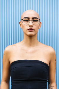 Confident young woman with shaved head wearing eyeglasses in front of blue wall