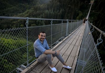 Portrait of man smiling while sitting on footbridge against forest