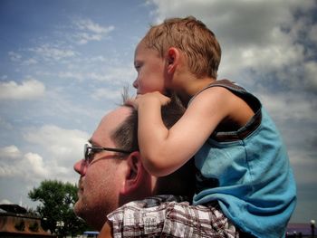 Father carrying son on shoulders against cloudy sky