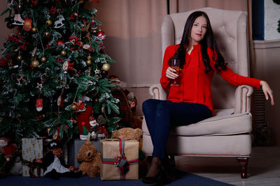 Portrait of young woman holding wineglass while sitting by christmas tree on armchair at home