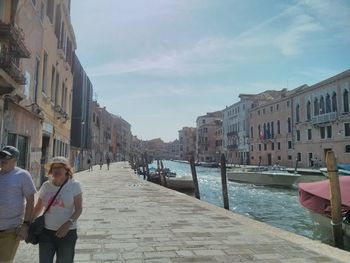 People on canal amidst buildings in city against sky