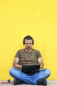 Young man using mobile phone while sitting against yellow wall