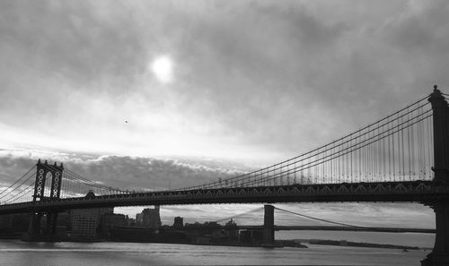 Low angle view of manhattan bridge over river against cloudy sky on sunny day