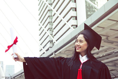 Smiling young woman holding graduation certificate while standing in city