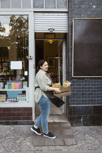 Side view of female owner standing outside deli holding crate