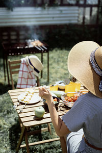 Young woman in summer hat sitting at the table, eating grilled vegetebles outdoors, rear view