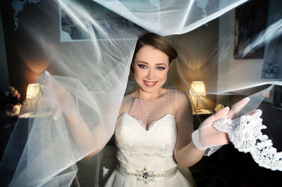 Portrait of happy beautiful bride standing amidst veil at home