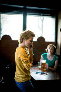 Young woman talking on phone against friend holding beer in restaurant