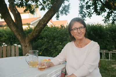 Portrait of smiling woman holding drink while sitting at table in backyard