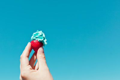 Cropped hand holding strawberry with whipped cream against clear blue sky