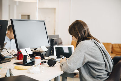 Side view of female programmer using mobile phone on desk while sitting in office