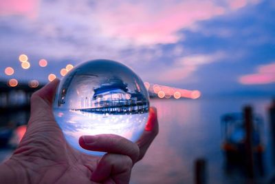 Cropped hand holding crystal ball by sea during sunset