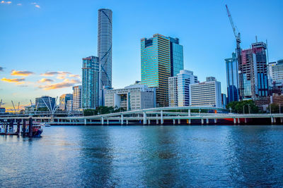 Modern buildings by brisbane river against sky during sunset