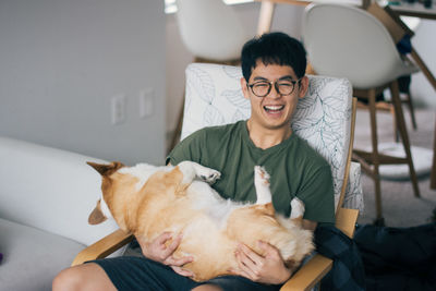 Portrait of happy young man holding dog while sitting on chair at home