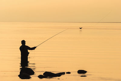 Silhouette man fishing in sea against orange sky