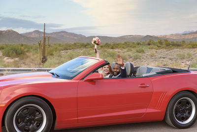 Portrait of happy newlywed couple with arms raised sitting in convertible at desert