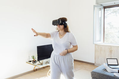 Young woman wearing virtual reality simulator gesturing in living room at home