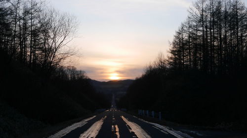 Road amidst silhouette trees against sky at sunset