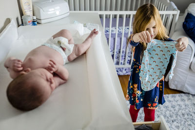 Girl choosing clothes for shirtless baby sister lying on table at home