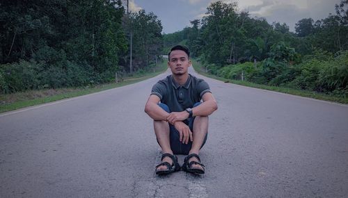 Portrait of young man sitting on road against trees