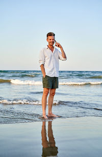 Full body barefoot male manager in white shirt and black shorts walking on beach near sea waves on a phone call on smartphone during summer vacation