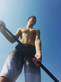 Low angle view of shirtless teenage boy holding oar against clear blue sky