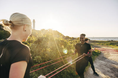 Smiling young man pulling resistance band while exercising with woman during team training session at beach