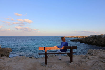 Woman sitting on bench looking at sea shore against sky