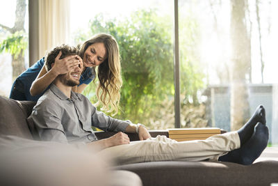 Smiling woman covering her boyfriend's eyes on sofa at home