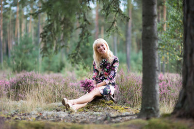 Portrait of woman holding flowers while sitting by tree trunk in forest