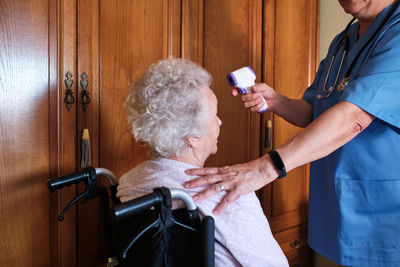 Side view of crop anonymous female nurse with infrared thermometer measuring body temperature of aged disabled female patient in wheelchair at home