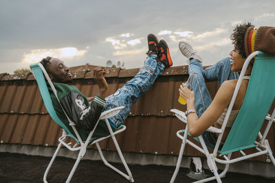 Full length cheerful male and female friends talking while sitting on chair at rooftop