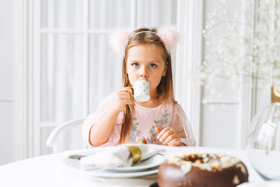 Funny cute little girl in light pink dress drinking tea on festive table in living room at home