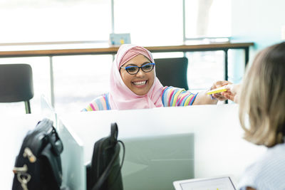 Positive muslim female in hijab sitting at table and giving paper stickers to unrecognizable coworker during work in office in costa rica in daytime
