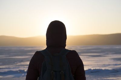 Rear view of man wearing hood while looking at sea against sky during sunset