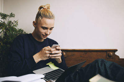 Distracted teenage boy with laptop texting on mobile phone while sitting at home