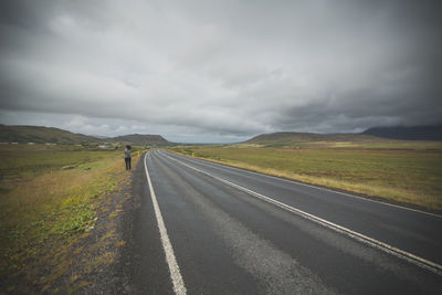Rear view of woman standing by empty road against cloudy sky